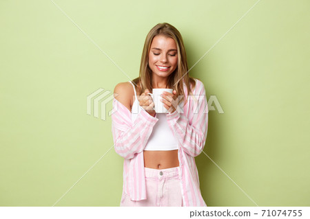 Image of beautiful tender woman with short blond hair, looking dreamy at cup, drinking coffee and smiling, standing over green background 71074755