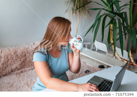 Middle age woman drinking tea sitting at the table at home working using computer laptop. Work from home during coronovirus pandemic concept Middle age woman drinking tea sitting at the table at home working using computer laptop. Work from home during coronovirus pandemic concept 71075394