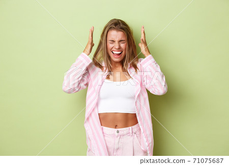 Potrait of distressed blond woman in pink shirt, holding hands on head and screaming, having depression, standing over green background 71075687
