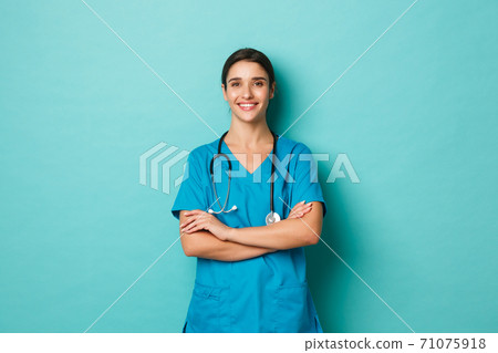 Coronavirus, pandemic and social distancing concept. Image of confident female doctor smiling, wearing scrubs with stethoscope, holding hands crossed on chest, posing over blue background 71075918