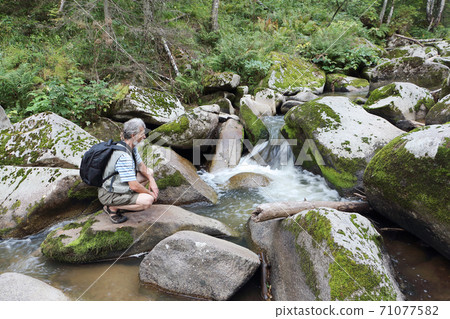 Man sitting on a stone near a mountain river Man sitting on a stone near a mountain river 71077582