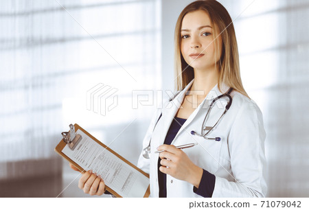 Young woman-doctor is making some notes using a clipboard, while standing in her cabinet in a clinic. Portrait of beautiful female physician with a stethoscope. Perfect medical service in a hospital 71079042