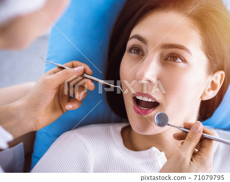 Smiling brunette woman being examined by dentist at sunny dental clinic. Hands of a doctor holding dental instruments near patient's mouth. Healthy teeth and medicine concept Smiling brunette woman being examined by dentist at sunny dental clinic. Hands of a doctor holding dental instruments near patient's mouth. Healthy teeth and medicine concept 71079795