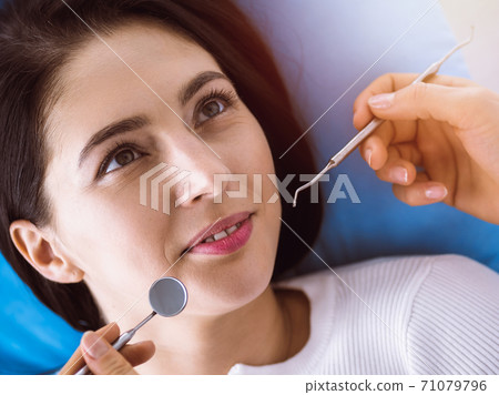 Smiling brunette woman being examined by dentist at sunny dental clinic. Hands of a doctor holding dental instruments near patient's mouth. Healthy teeth and medicine concept 71079796