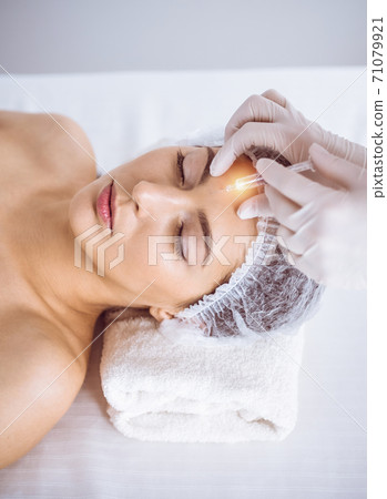 Beautician doing beauty procedure with syringe to face of young brunette woman in sunny spa center. Cosmetic medicine and surgery, beauty injections Beautician doing beauty procedure with syringe to face of young brunette woman in sunny spa center. Cosmetic medicine and surgery, beauty injections 71079921