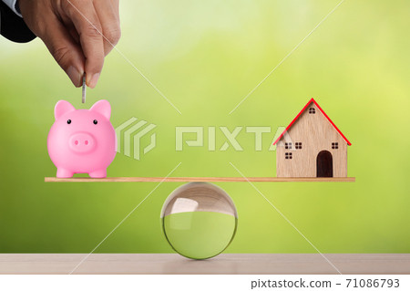 Businessman hand putting coin into piggy bank with model house on wooden marble seesaw balancing on green background. Property investment and home mortgage financial real estate advertising concept Businessman hand putting coin into piggy bank with model house on wooden marble seesaw balancing on green background. Property investment and home mortgage financial real estate advertising concept 71086793