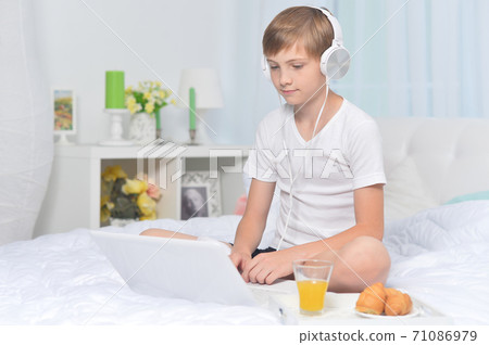 Boy listening to music on bed while having breakfast Boy listening to music on bed while having breakfast 71086979