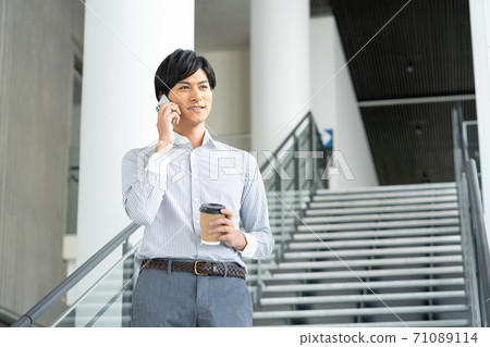 A smiling young man having a conversation on his smartphone while having a drink in the office lobby A smiling young man having a conversation on his smartphone while having a drink in the office lobby 71089114