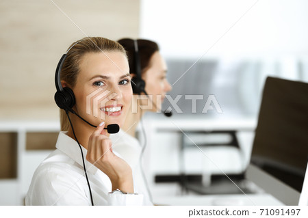 Blond female customer service representative and her colleague are consulting clients online using headset. Call center and business people concept Blond female customer service representative and her colleague are consulting clients online using headset. Call center and business people concept 71091497