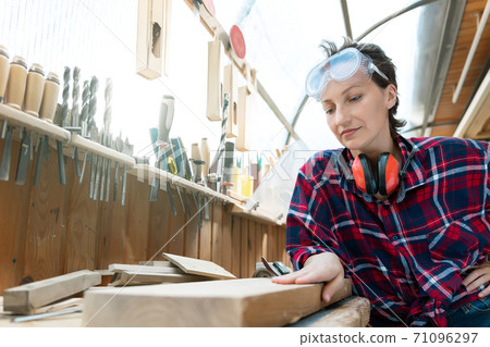 Young beautiful handy professional happy female strong carpenter portrait wearing protective goggles working in carpentry diy workshop against wood. Confident engineer. Women male hobby at workbench 71096297