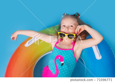 Funny little girl dressed in a swimsuit and sunglasses lying on an inflatable color ring on blue studio background. Funny little girl dressed in a swimsuit and sunglasses lying on an inflatable color ring on blue studio background. 71096771