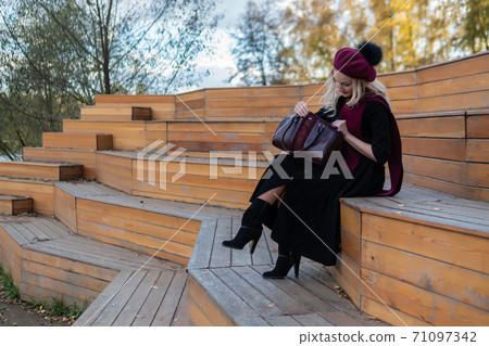 A beautiful woman sits on a wooden summer theater bench in a burgundy coat and biret, looks at the camera, in the fall against the blue sky. 71097342