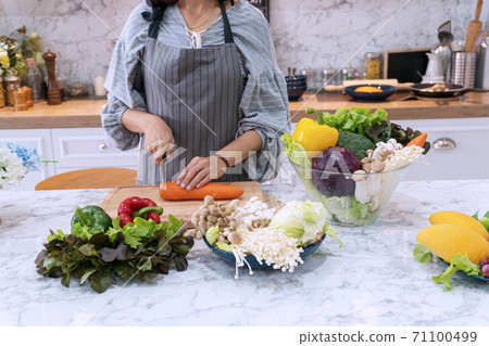 Attractive woman holding carrot with kitchen knife on cutting board in kitchen. 71100499