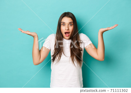 Surprised attractive woman in white t-shirt, raising hands up and gasping amazed, standing over blue background 71111302