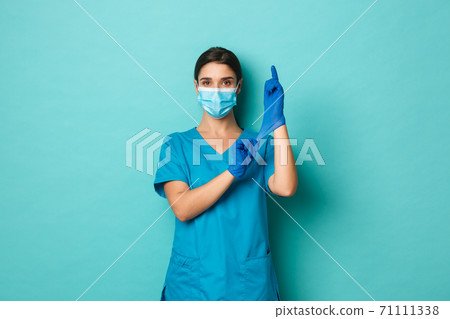 Covid-19, pandemic and medicine concept. Confident beautiful woman in medical mask and crubs, put on gloves and looking self-assured, getting ready for patient check up Covid-19, pandemic and medicine concept. Confident beautiful woman in medical mask and crubs, put on gloves and looking self-assured, getting ready for patient check up 71111338