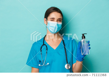 Concept of covid-19 and quarantine concept. Close-up of beautiful female doctor in medical mask, gloves and scrubs, winking and showing hand sanitizer, standing over blue background 71111368