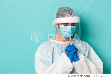 Concept of covid-19, pandemic and quarantine. Close-up of hopeful female doctor in personal protective equipment, holding hands in pray, begging or pleading, standing over blue background 71112120
