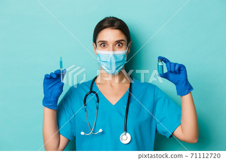Concept of covid-19 and quarantine concept. Close-up of female doctor in medical mask, latex gloves and scrubs holding syringe and showing coronavirus vaccine, looking amazed 71112720