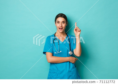 Coronavirus, pandemic and social distancing concept. Image of excited female doctor, wearing scrubs, having an idea, raising finger up in eureka gesture, standing over blue background 71112772