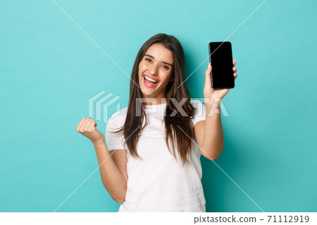 Portrait of successful and happy woman, showing smartphone screen and making fist pump gesture to celebrate win, achieve goal in application, standing over blue background 71112919