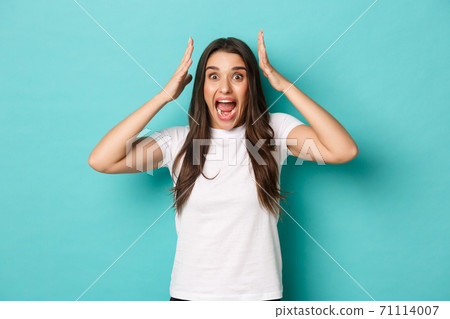 Image of scared and alarmed brunette girl in white t-shirt, shouting and holding hands over head, standing in panic over blue background 71114007