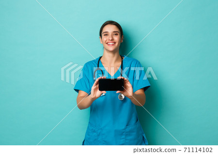 Coronavirus, pandemic and social distancing concept. Image of smiling female doctor in scrubs, showing smartphone horizontally, standing over blue background Coronavirus, pandemic and social distancing concept. Image of smiling female doctor in scrubs, showing smartphone horizontally, standing over blue background 71114102