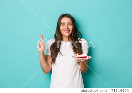 Image of happy and excited brunette woman, celebrating her birthday, close eyes and cross fingers while making wish, holding b-day cake with lit candle 71114103