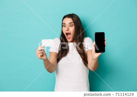 Portrait of fascinated brunette girl, showing mobile phone screen and looking excited at credit card, standing against blue background Portrait of fascinated brunette girl, showing mobile phone screen and looking excited at credit card, standing against blue background 71114129