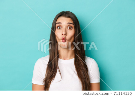 Close-up of silly brunette woman, sucking lips and showing funny faces, standing over blue background Close-up of silly brunette woman, sucking lips and showing funny faces, standing over blue background 71114130
