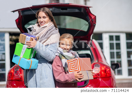 Happy mom and daughter standing back to back with gift boxes at car trunk 71114424