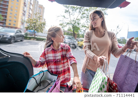 Girl and young female looking at each other, taking out shopping bags from car trunk Girl and young female looking at each other, taking out shopping bags from car trunk 71114441