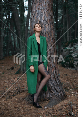 Fashionable young woman in green coat posing in a forest near the tree Fashionable young woman in green coat posing in a forest near the tree 71119712