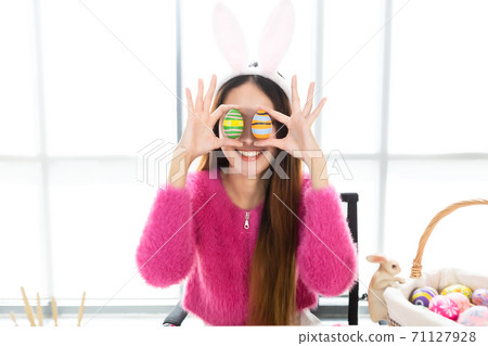 Happy smile Asian Young woman wearing bunny ears and holding up a colorful Easter egg in front of her eye Skewers In the white room background 71127928