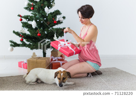 Happy woman with dog opening Christmas gifts. Christmas tree with presents under it. Decorated living room 71128220
