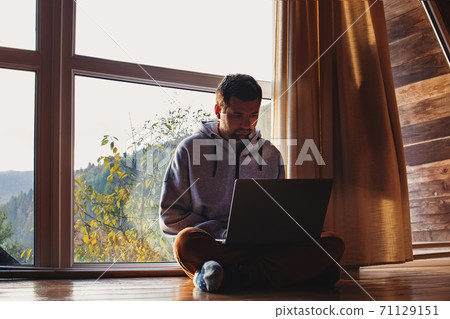 Man using laptop sitting near big window in wooden cottage 71129151