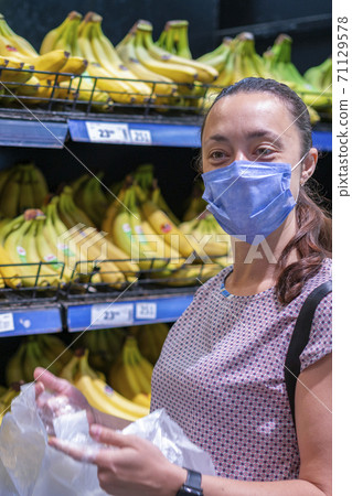 Focused woman in face mask choosing fruits, taking bananas from shelves in grocery store. Customer in supermarket. Side view. Shopping during epidemic concept. vertical photo Focused woman in face mask choosing fruits, taking bananas from shelves in grocery store. Customer in supermarket. Side view. Shopping during epidemic concept. vertical photo 71129578