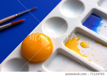 White palette with watercolor paints and brushes. Blue background. Instead of yellow, the egg yolk is used. Creative design and art. Top view and copy space 71129783