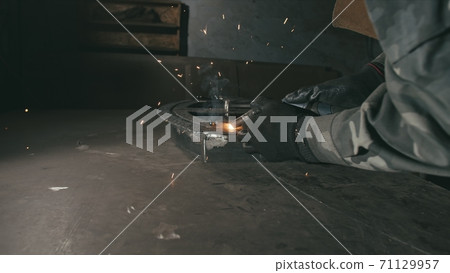 Forge workshop. Smithy. Worker in a welding hood helmet welds a part by electric welding. Sparks are reflected in the protective screen. 71129957