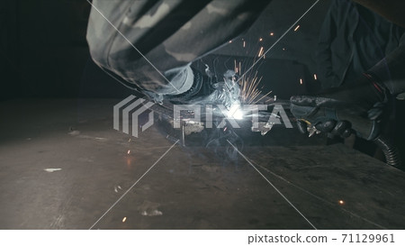 Forge workshop. Smithy. Worker in a welding hood helmet welds a part by electric welding. Sparks are reflected in the protective screen. Forge workshop. Smithy. Worker in a welding hood helmet welds a part by electric welding. Sparks are reflected in the protective screen. 71129961