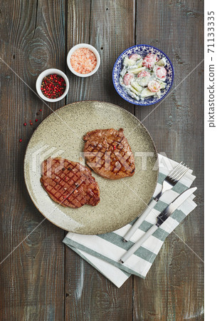 Two grilled ribeye steaks, spices and tomato-cucumber salad with sour cream on a wooden table. Two grilled ribeye steaks, spices and tomato-cucumber salad with sour cream on a wooden table. 71133335