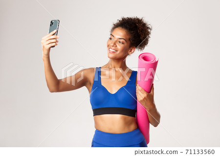 Young beautiful african-american fitness woman in sport outfit, taking selfie with yoga mat, taking photo on smartphone, standing against white background Young beautiful african-american fitness woman in sport outfit, taking selfie with yoga mat, taking photo on smartphone, standing against white background 71133560