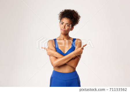 Concept of workout and fitness. Sad indecisive african-american girl in sports clothing, looking confused, pointing fingers sideways, white background 71133562