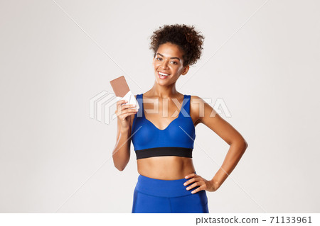 Young athletic woman in blue sportswear looking happy, holding chocolate bar and smiling pleased, standing against white background 71133961