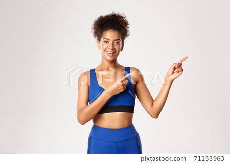 Concept of workout and fitness. Attractive fit african-american female athlete in blue sports costume, pointing fingers right and smiling, showing advertisement, white background 71133963