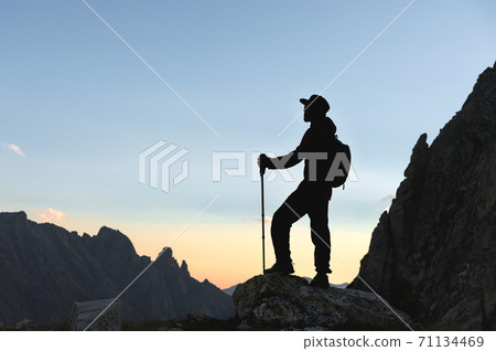 Silhouette of a man traveler in a cap with a backpack and trekking poles. Stands on the edge of a cliff against the background of mountains and the skyline in the evening at dusk 71134469