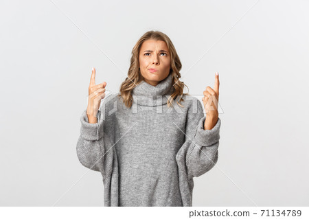 Image of skeptical blond girl in grey sweater, looking indecisive and pointing up, smirking doubtful, standing over white background 71134789