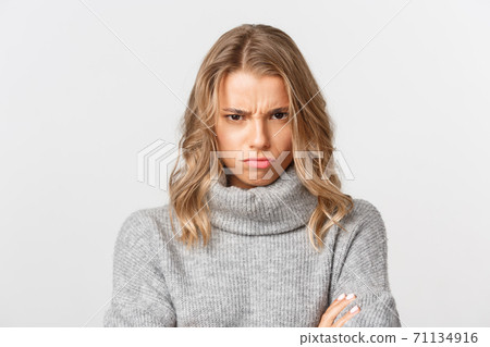 Close-up of angry blond girl in grey sweater, looking from under forehead and sulking, feeling offended, standing over white background Close-up of angry blond girl in grey sweater, looking from under forehead and sulking, feeling offended, standing over white background 71134916