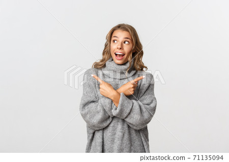 Amazed blond girl pointing fingers sideways, have two choices but looking happy at upper left corner, standing over white background 71135094