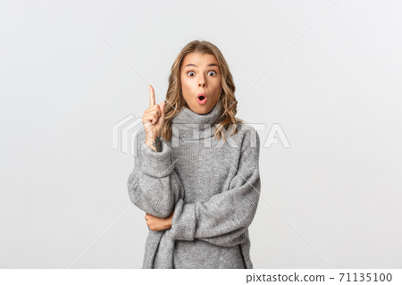 Close-up of thoughtful cute girl in grey sweater, having an idea, raising finger and say something, standing over white background 71135100