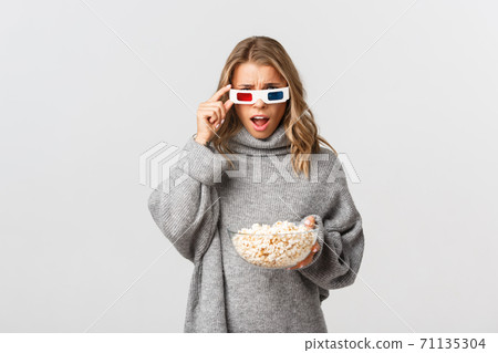 Image of blond girl in 3d glasses, holding bowl with popcorn, watching movie and looking confused, standing over white background Image of blond girl in 3d glasses, holding bowl with popcorn, watching movie and looking confused, standing over white background 71135304